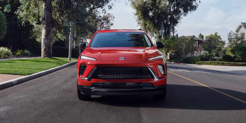 A vibrant red SUV Envision drives down a sunny suburban street. Its sleek design and bold grille convey modernity, surrounded by lush greenery and houses.