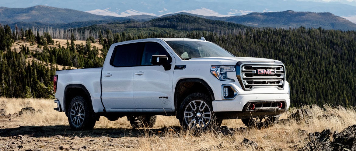A white GMC pickup truck Sierra 1500 AT4 with rugged tires is parked on a grassy hill overlooking a vast forest and distant mountains under a clear blue sky.
